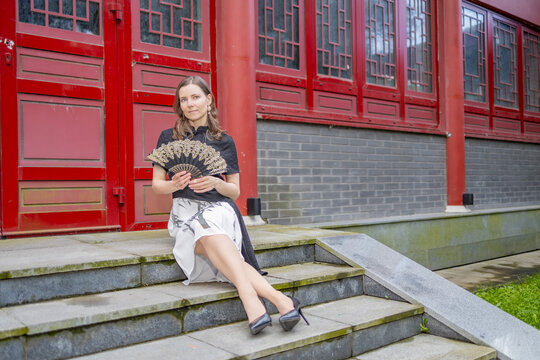 Stylish woman in a black and white dress holding a decorative fan while sitting gracefully on stone stairs in front of a red architectural facade - Powered by Adobe