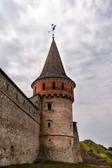 Medieval Kamianets-Podilskyi fortress tower with stone walls and a conical roof, surrounded by lush greenery and dramatic clouds, showcasing historical architecture and cultural heritage