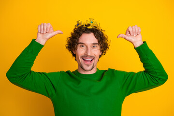 Cheerful young man in a green sweater wearing a crown playfully pointing thumbs at himself on a...