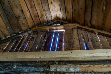 Close up view of an old wooden attic wall showing weathered planks with visible gaps. Daylight streams through the cracks, revealing the aged texture and rustic construction of the structure.