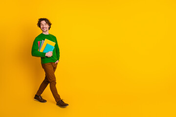 Young cheerful curly-haired man in green sweater with stationery on yellow backdrop depicting...