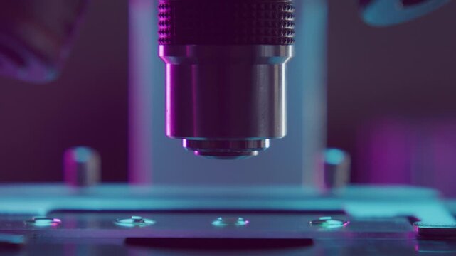 Detailed close up view of microscope objective lens focusing above specimen slide during examination in laboratory with vivid purple and blue lighting