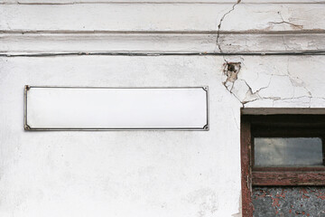 Old damaged building wall with cracked plaster and empty signboard above window. Weathered facade with peeling paint and broken surface texture. Urban decay architecture background with copy space.