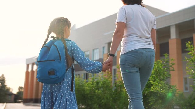 Parent walk with child holding hand near school building under warm sunlight, child wearing blue backpack and polka dot dress, adult in jeans guiding child along green path toward entrance smile