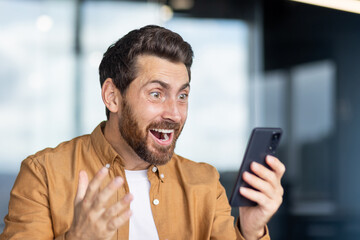 Excited man reacts to smartphone message with amazement and joy indoors.