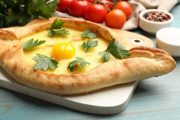 Tasty khachapuri with cheese, egg, parsley, tomatoes and spices on light blue wooden table, closeup