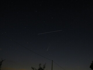 The night sky illuminated by distant stars, featuring Comet NEOWISE with its visible tail and a bright satellite light trail, over a dark horizon with faint light pollution and power lines.