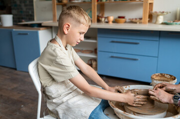 Boy and female instructor shaping clay together at pottery wheel, kids learning traditional craft