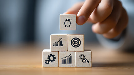 Hand placing wooden cube with alarm clock symbol atop stacked business icons wooden cubes strategy