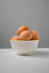 A white ceramic bowl filled with brown eggs on a white table against a gray backdrop; soft light, minimal still life ideal for grocery catalogs.