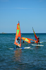 The sea and people windsurfing. They are standing on boards with bright red and orange-yellow sails. The water is calm and the sky is bright blue, with one small cloud