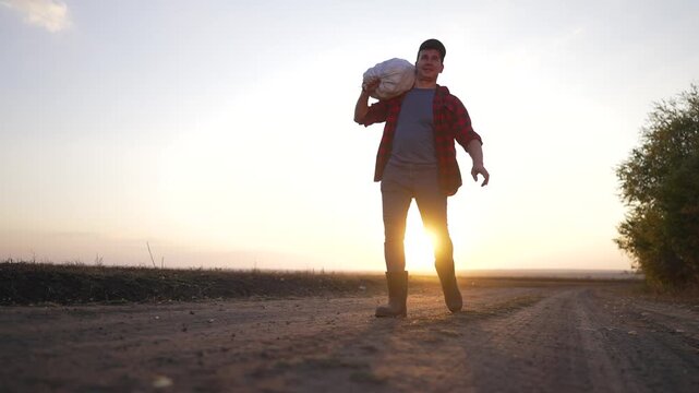 Man walking on dirt road carrying sack while sun sets behind field horizon with boot and cap wearing flannel shirt showing farmer determination and rural work at sunset warm glow and long shadow
