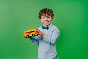 Happy preschool boy wearing bow tie playing with school bus toy over green background