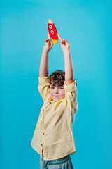 Little boy holding paper rocket toy standing over blue background