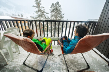 Two children face each other in deck chairs on snowy balcony
