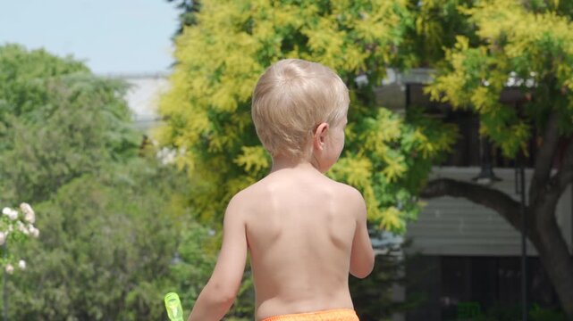 A young boy is blowing bubbles with a bubble wand. He is outdoors in a lush garden with bright green and yellow foliage on a sunny day.