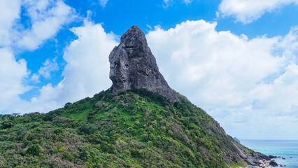 O Morro do Pico em Fernando de Noronha visto de cima, com destaque para sua formação rochosa e a vegetação ao redor, além da costa azul cristalina, cenário perfeito para fotos aéreas, turismo