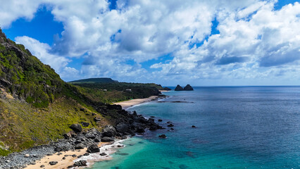 Aérea de praia e costa íngreme em Fernando de Noronha, destacando mar azul intenso, formações rochosas e o Morro do Pico em paisagem tropical preservada