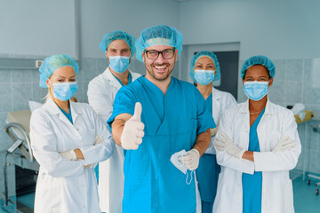 Fototapeta premium Confident Male Surgeon and Diverse Medical Team in Bright Hospital Operating Room Giving Thumbs Up