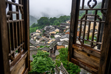 Through an ornate window frame, white-walled buildings with black-tiled roof and upturned eaves are classic Huizhou architecture veiled in mist at Huangling Village in Wuyuan, Jiangxi, China