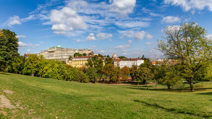 Fototapeta premium Sunny autumn panorama of a beautiful park in Buda with wide green lawn surrounded by yellow-green trees and view of historic Buda Castle against blue sky. Budapest, Hungary.
