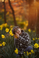 A child, wearing glasses and a plaid coat, sits among yellow flowers holding one cheek in a sunlit...