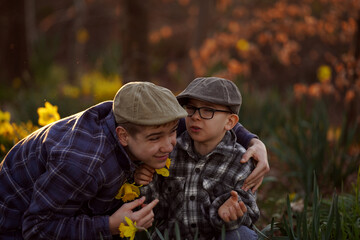 Two boys in plaid shirts and hats are sitting among yellow flowers, with one embracing the other....