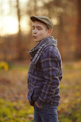 Young person in plaid jacket and cap poses outdoors at sunset with hands in pockets, giving a...