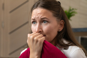 Stressed young woman gripping a red pillow and anxiously biting her fingers, surrounded by a tense...
