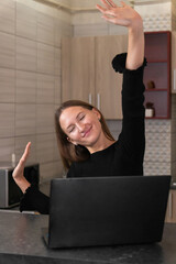 Young woman taking a break from working on her laptop in the kitchen, stretching her arms and...