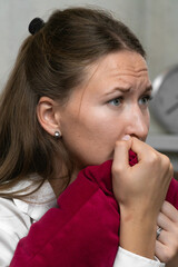 Stressed young woman gripping a red pillow and anxiously biting her fingers, surrounded by a tense...