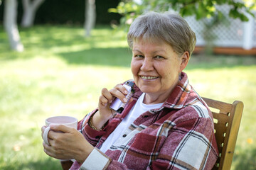 Smiling elderly woman 60-70 years in red plaid shirt and white t-shirt sitting outdoors on sunny...