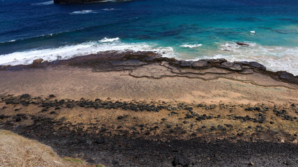Imagem aérea da orla da Praia do Leão em Noronha, com ondas suaves, areia dourada e falésias cobertas por vegetação tropical, refletindo a beleza natural do arquipélago