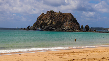 Formação rochosa diante do mar calmo em Fernando de Noronha, vista a partir da faixa de areia. A cena combina tons naturais e destaca a beleza do litoral preservado do arquipélago