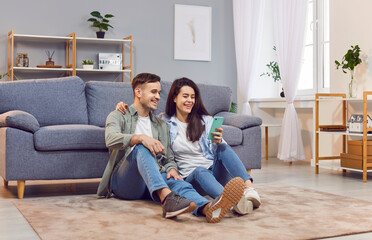 Young happy couple sitting on floor near sofa, using mobile phone together for online...