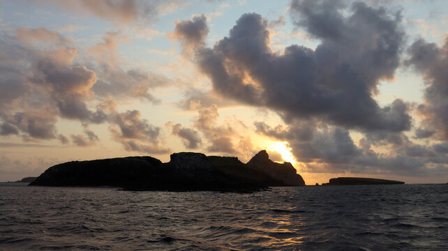P&ocirc;r do sol em Fernando de Noronha com silhueta de ilhas no mar, c&eacute;u dram&aacute;tico, nuvens iluminadas e reflexos dourados na &aacute;gua, criando paisagem tropical e contemplativa.