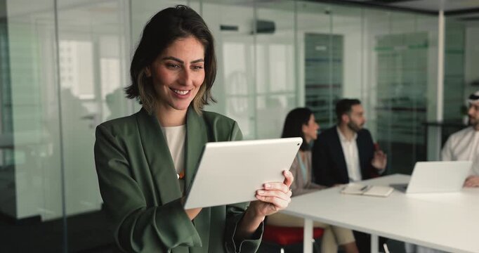 Smiling Mexican businesswoman stands in meeting room using modern tablet device, working on data or reports, reviewing documents, editing proposals, managing project objectives and details. Tech usage