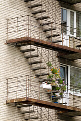 Exterior view of an old brick building with weathered fire escapes, decorated by colorful potted plants and flowers on a middle landing.