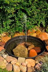 źródełko w ogrodzie, mini fontanna solarna otoczona kamieniami i zielonymi roślinami, tryskający strumień wody z małej fontanny ogrodowej, a solar fountain in a small bowl in the garden  © kateej