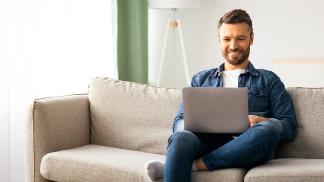Handsome bearded man using laptop and smiling, sitting on couch in living room, websurfing or using social media, copy space. Cheerful middle-aged man enjoying part-time remote job