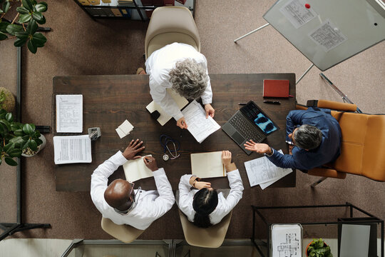Group of middle aged and senior multiethnic doctors and one middle aged Caucasian man sitting around table discussing medical documents and using laptop during meeting, top view
