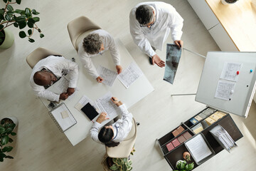 Group of middle aged and senior multiethnic doctors discussing medical documents and analyzing X-ray image around table in modern office, top view showing collaborative teamwork