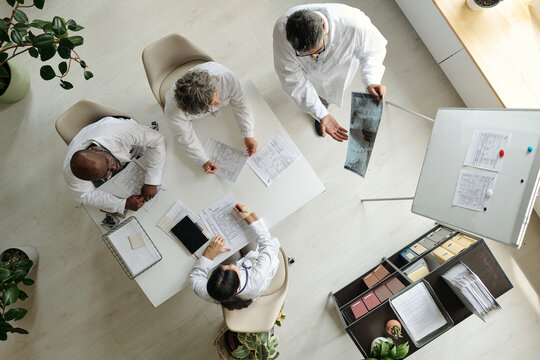 Group of middle aged and senior multiethnic doctors discussing medical documents and analyzing X-ray image around table in modern office, top view showing collaborative teamwork