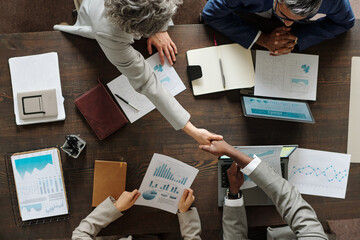 Top view of middle aged Caucasian woman and Black man shaking hands over table during business meeting with diverse colleagues