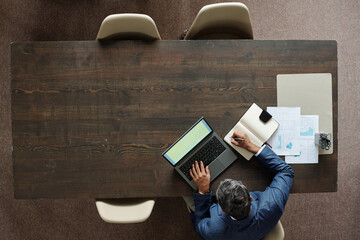 Middle aged Caucasian man working at desk using laptop and writing in notebook, analyzing documents with charts and graphs, top view showing workspace and office supplies