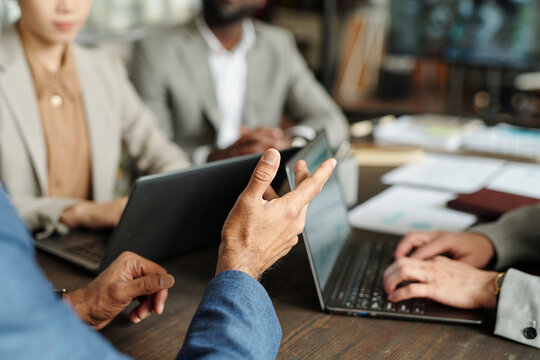 Multiethnic group of middle aged professionals collaborating around laptops during business meeting, hands gesturing while discussing project, focus on teamwork and communication