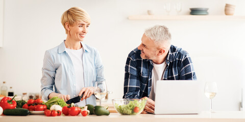 Free time together, preparing food for dinner and work remotely at home. Smiling senior woman cuts vegetables, husband looks at her in kitchen with vegetables, laptop and glasses of wine on table