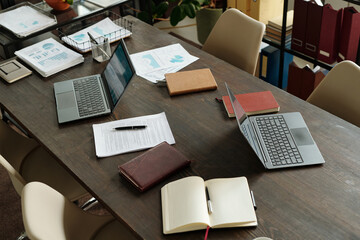 Top view showing two open laptops, scattered documents, notebooks, and pen on wooden meeting table,...
