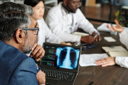 Middle aged Caucasian man reviewing chest X-ray image on laptop with Asian woman and Black man in medical uniforms discussing diagnosis at table during meeting, hands gesturing in conversation