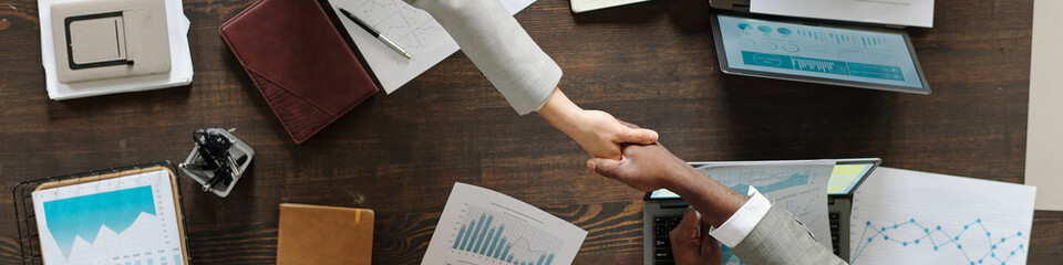 Caucasian woman and Black man shaking hands over wooden desk with financial charts and laptops...